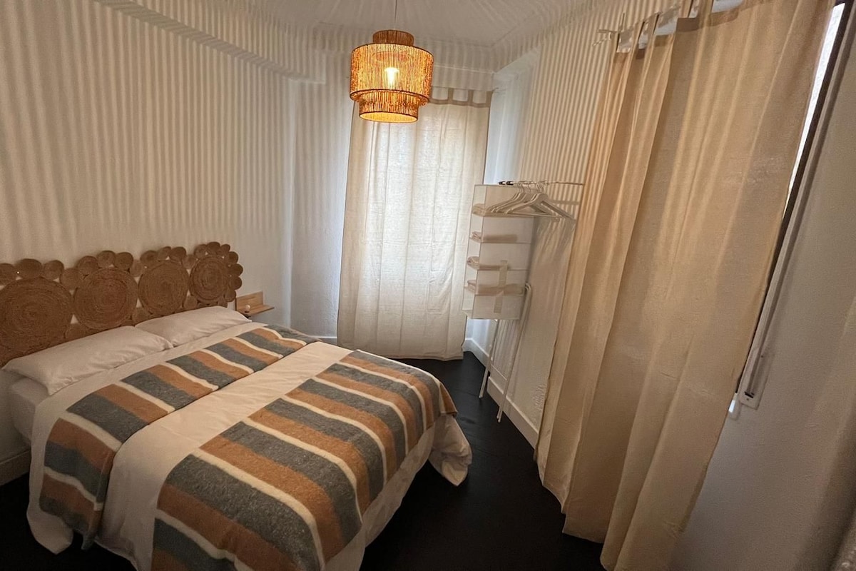 A hotel room with a bed, a hanging light fixture, a coat rack, and a window with curtains.