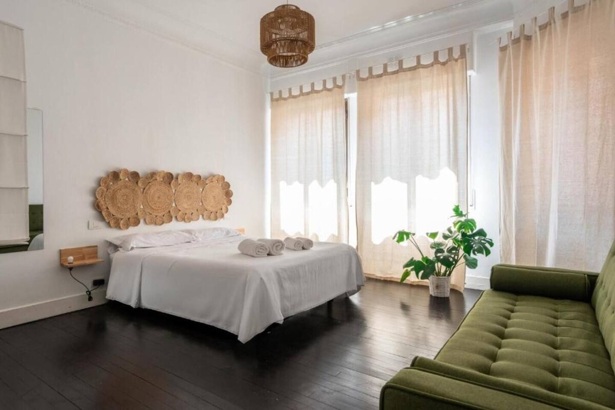 A bedroom with a bed, a green sofa, a potted plant, and a wall-mounted light fixture.