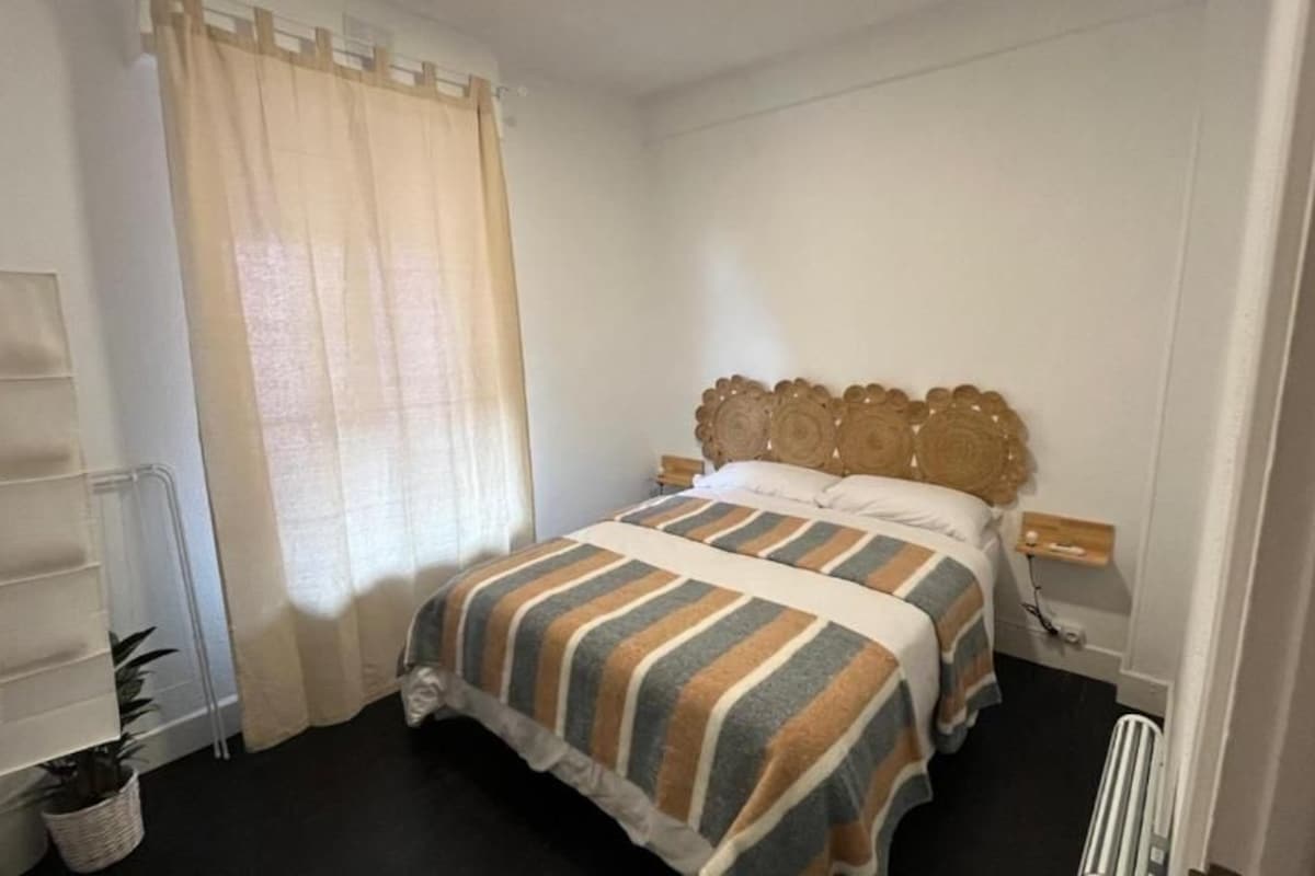 A bedroom with a bed, a bedside table, a nightstand, a window with curtains, and a radiator.