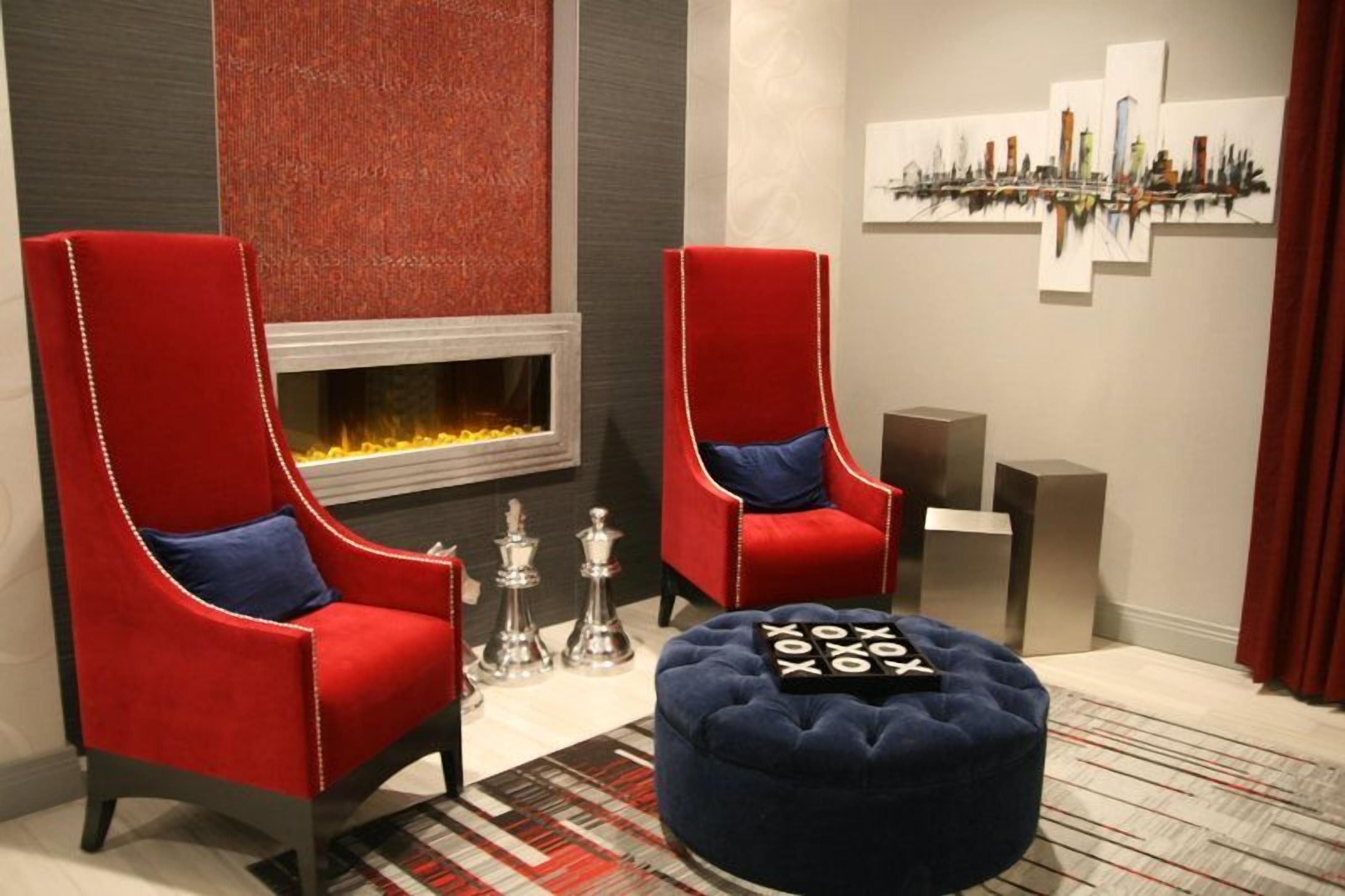 A modern lounge area with two red upholstered chairs, a blue ottoman, a fireplace, and a cityscape painting on the wall.