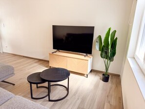 Ett modernt vardagsrum med en platt-TV, ett träskåp, ett runt bord, en krukväxt och en soffa.