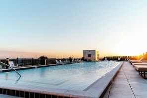 Ein Pool auf dem Dach mit Liegestühlen und ein kleines Gebäude, vermutlich eine Umkleide oder ein Stauraum, unter blauem Himmel.