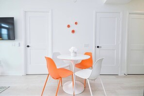 Et værelse med et rundt, hvidt bord, orange og hvide stole og en hvid vase med blomster. Der er hvide døre og et fladskærms-tv monteret på væggen.