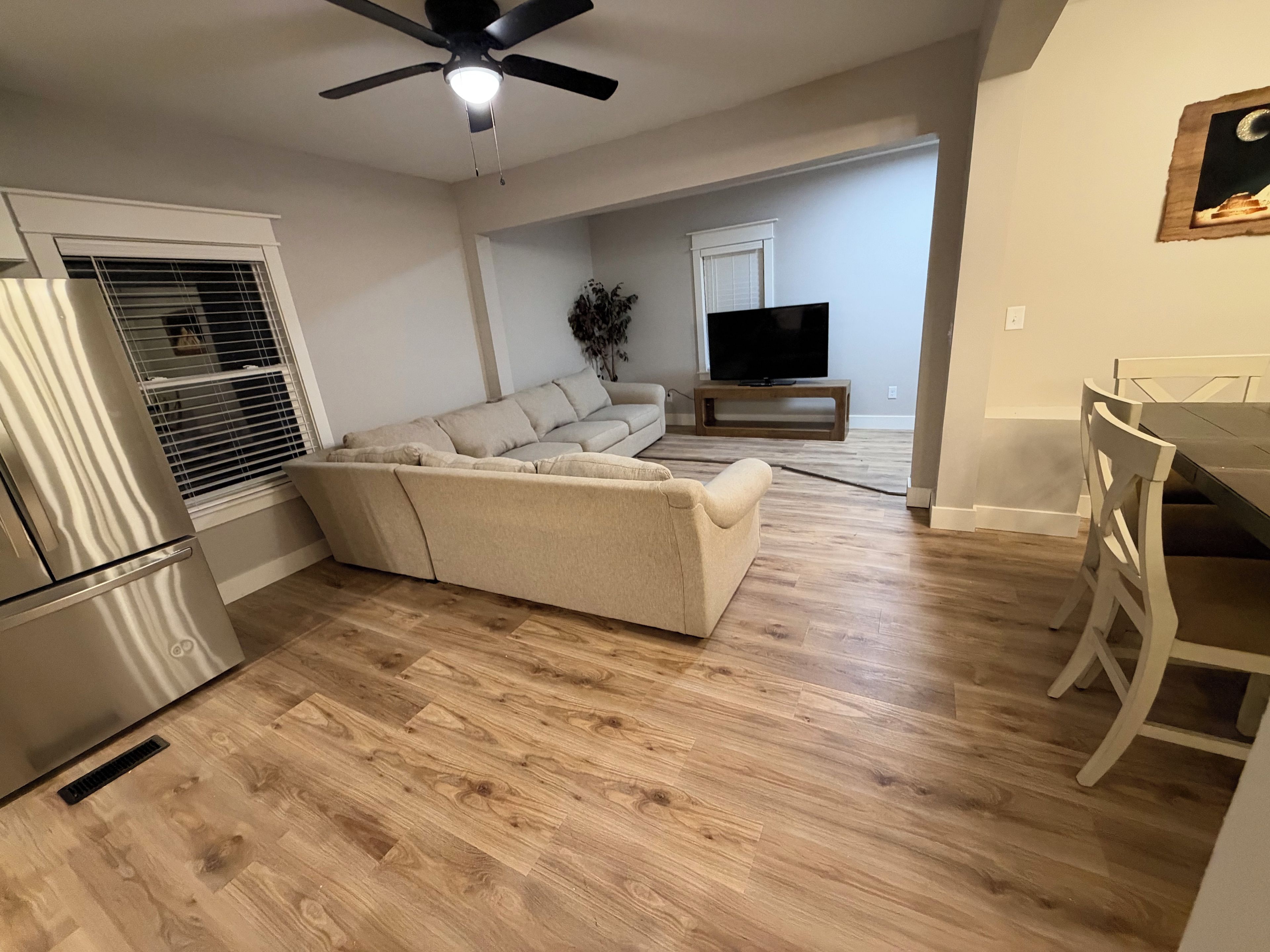 A living room with a sectional sofa, a flat-screen TV on a wooden stand, a ceiling fan, and a dining area with a table and chairs.