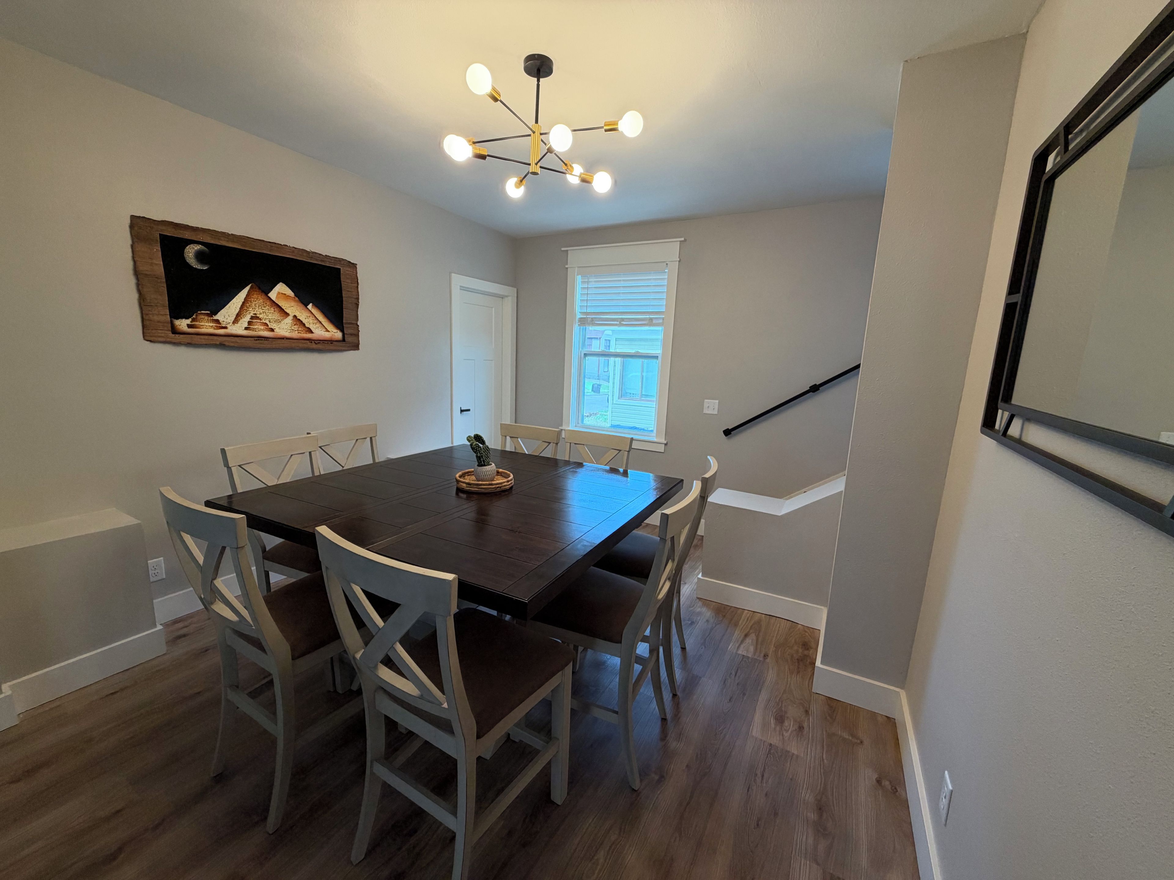 A dining room with a dark wooden table and six chairs, a chandelier, a framed picture on the wall, and a window with blinds.
