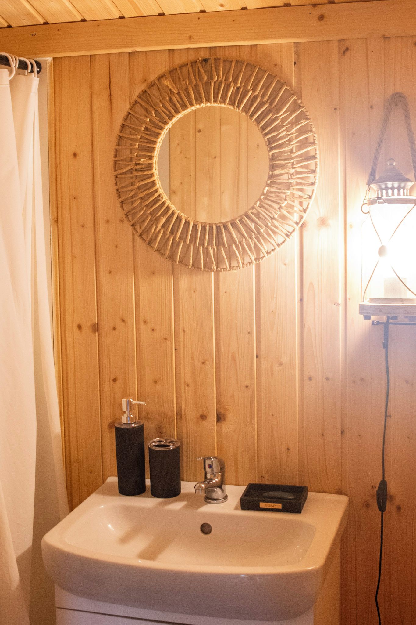 Ein Badezimmer mit einer Holzwand, einem runden Spiegel, einem Waschbecken mit Seifenspender und einem Handtuchhalter.