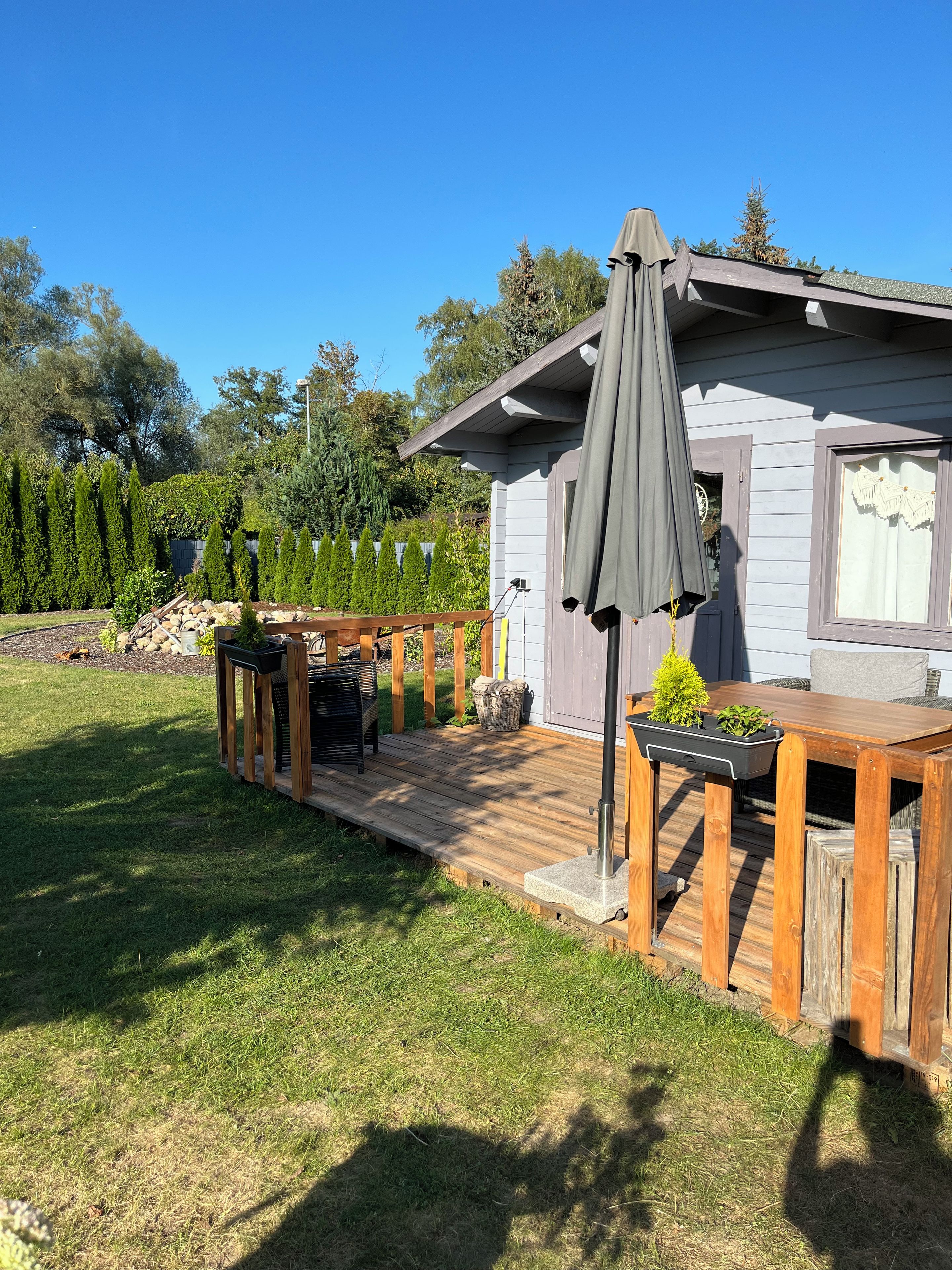 A wooden deck with a table and chairs, a pergola with a canopy, and a small garden area with trees and shrubs.