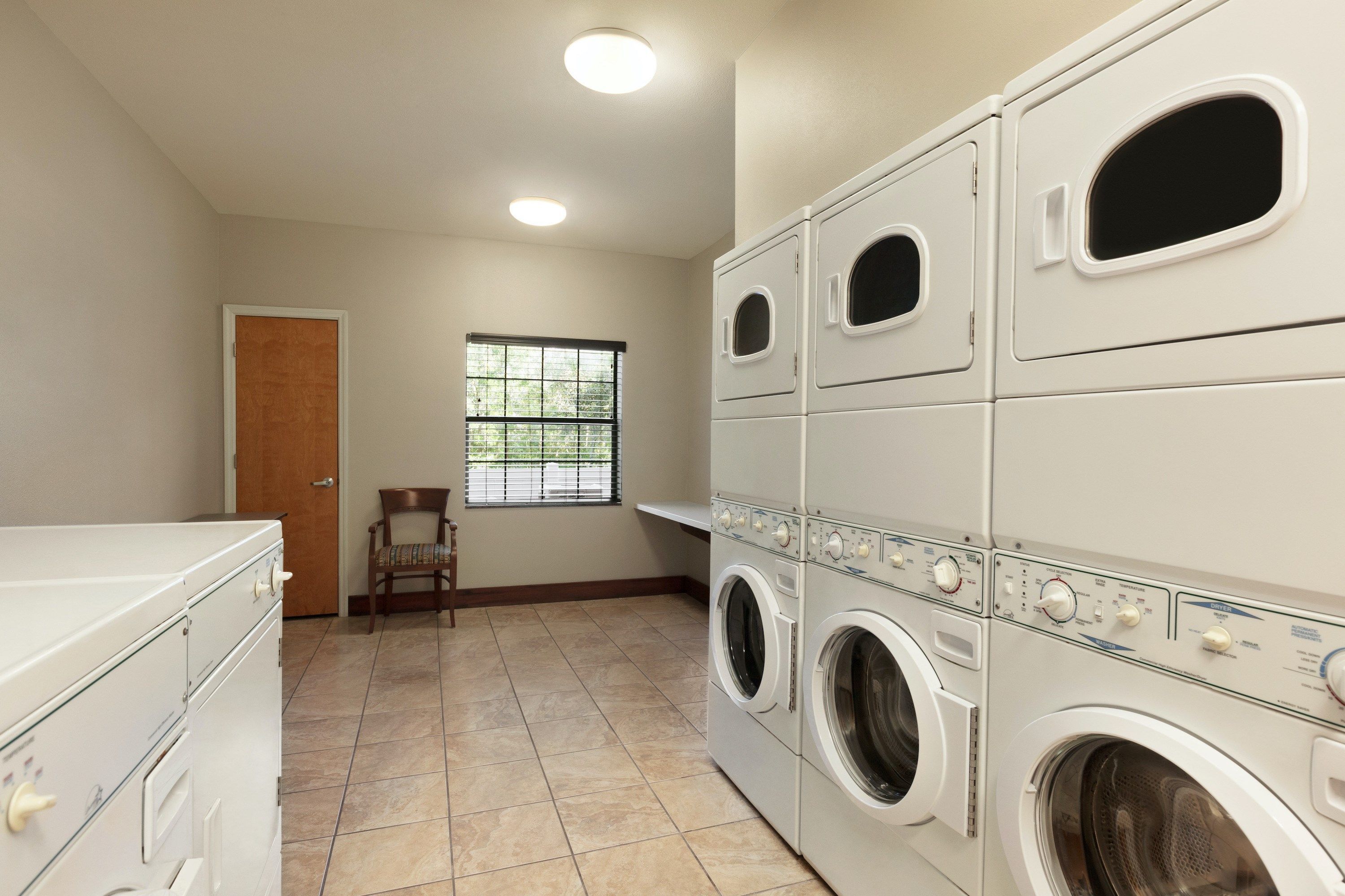 A laundry room with stacked washers and dryers, a wooden chair, a door, and a window with blinds.