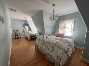 A bedroom with a sloped ceiling, a chandelier, a bed with floral bedding, a rocking chair, and a window with curtains. - The Gnome Home in McKennan Park (Sioux Falls)
