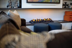 A living room with a sofa, ottomans, a fireplace with burning logs, and a television mounted on the wall. - AMB203 | Dog Friendly | New Luxury Complex | Hot Tub | Winter Shuttle (Steamboat Springs)
