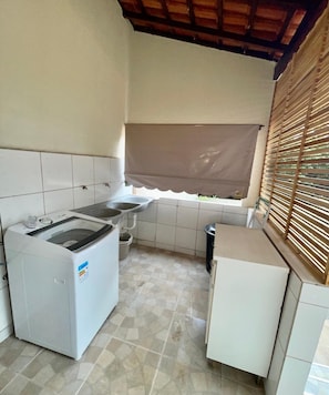 Una cocina compacta con lavadora, un refrigerador pequeño y un ventanal con persiana marrón.