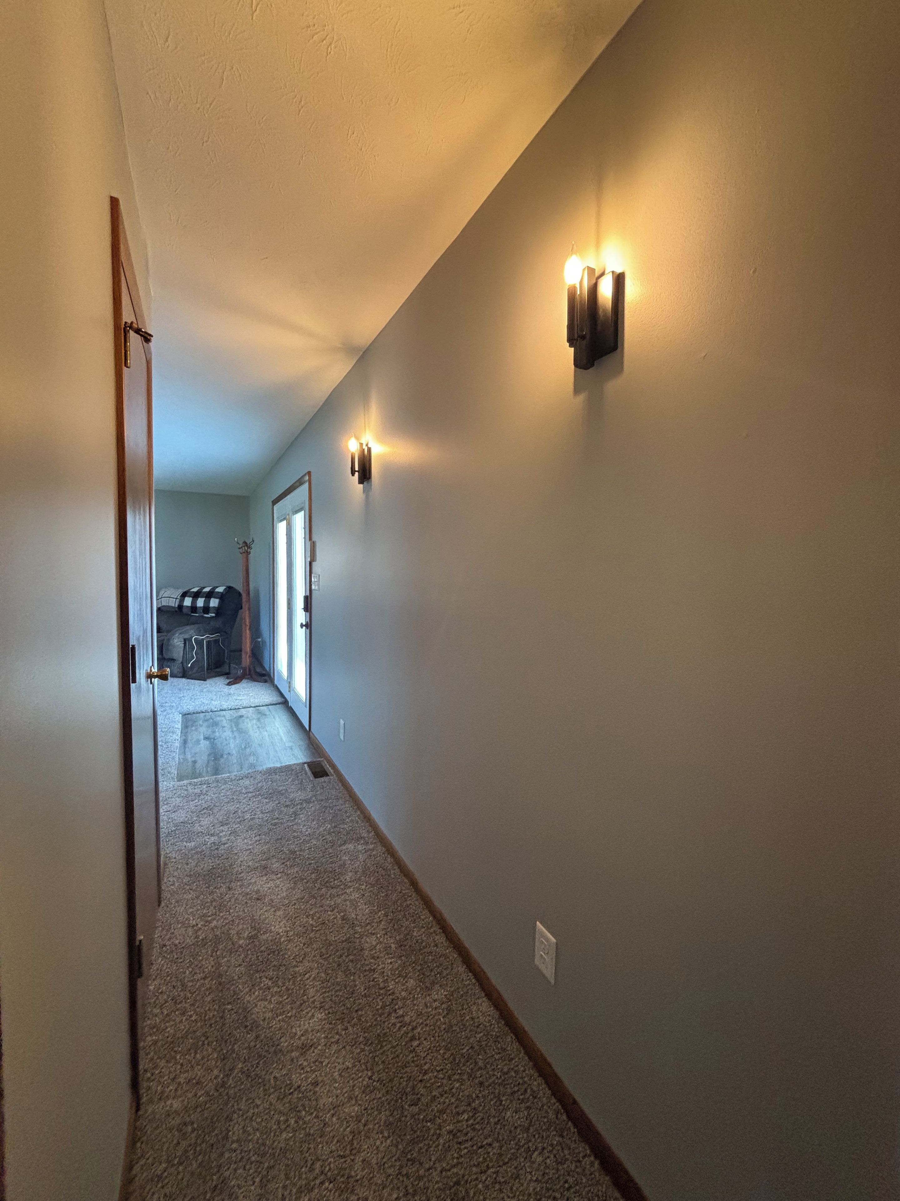 A hallway with carpeted flooring, a door on the left, and a light fixture on the wall.