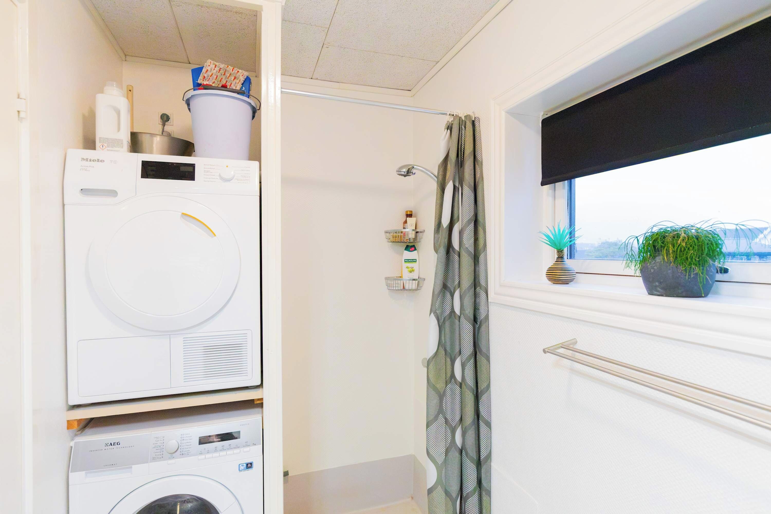 A laundry area with a front-loading washing machine and a dryer, a shower with a patterned curtain, and a window with a view.