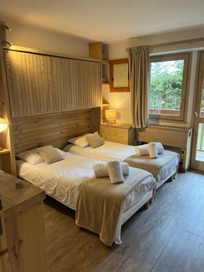 A hotel room with two beds, wooden headboard, bedside lamps, a window with curtains, and a view of greenery. - Sunny apartment, close to the slopes and shops (Les Allues)