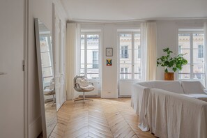 Ein Hotelzimmer mit Fischgrätparkett, einem weißen Bett, einem Sessel, einer Topfpflanze und Blick auf die Stadt durch große Fenster.