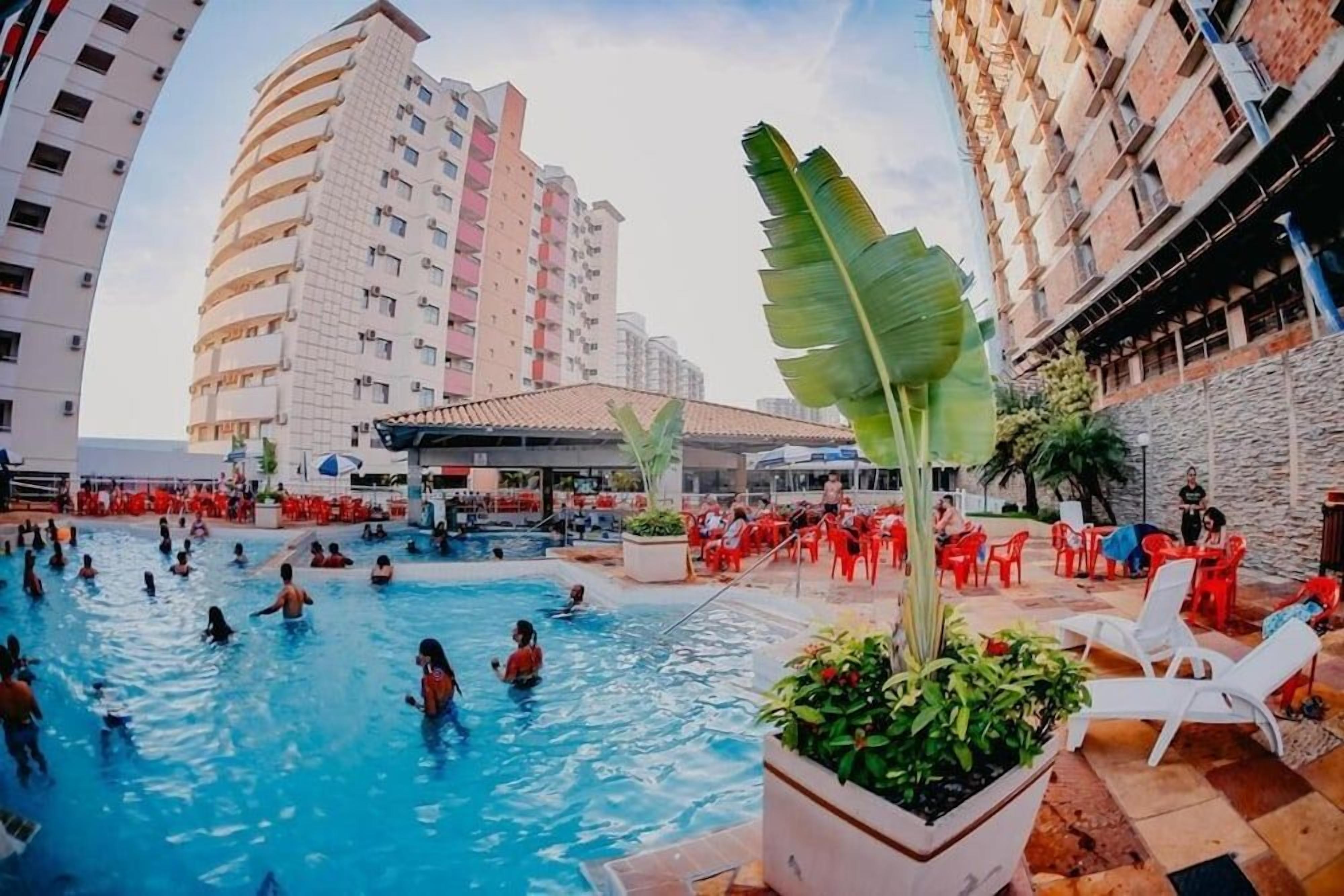A swimming pool area with people swimming, surrounded by red chairs and tables, with buildings in the background.