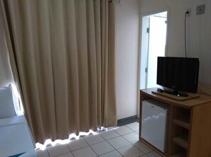 Ein Zimmer mit Fernseher auf Holzfuß, einer kleinen Kühlmöglichkeit und einem Fenster mit Vorhängen.