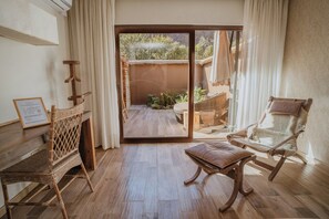 Una habitación con piso de madera, una silla de mimbre, un banco acolchado, un piano y vistas a un área exterior con plantas a través de un vidrio.