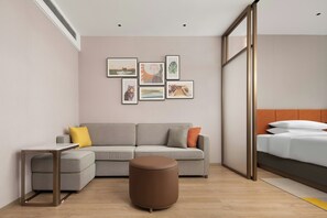Una habitación de hotel moderna con un sofá gris, una mesita auxiliar pequeña, un reposapiés marrón y una cama con ropa de cama blanca.