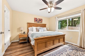 A bedroom with a wooden bed, a nightstand, a ceiling fan, a window with curtains, and a patterned rug. - Near UF & Downtown | Spacious Home + Backyard (Gainesville)