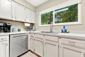A modern kitchen with white cabinets, a dishwasher, a coffee maker, a toaster, and a window with a view of trees. - Near UF & Downtown | Spacious Home + Backyard (Gainesville)