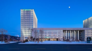 A modern hotel with a prominent entrance, a large parking lot, and a tall building in the background.