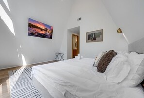 Een slaapkamer met een groot bed, een schilderij van een zonsondergang, een klein tafeltje en een deur naar een andere kamer.