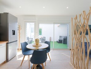 A modern kitchen with a built-in refrigerator, a dining area with a round table and chairs, and a view of a green outdoor space through glass doors. - Apartment with AC and spa in picturesque village (Portbail)