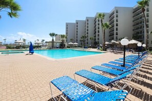 Un espace aménagé au bord de la piscine, avec des chaises longues et des parasols, entouré de palmiers et bordé d’un immeuble à plusieurs étages.