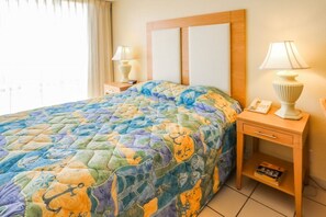 A hotel room with a large bed, a nightstand with a lamp, a bedside table with a phone, and a window with curtains. - Beachfront 1BR Condo w/ Balcony View + King Bed (South Padre Island)