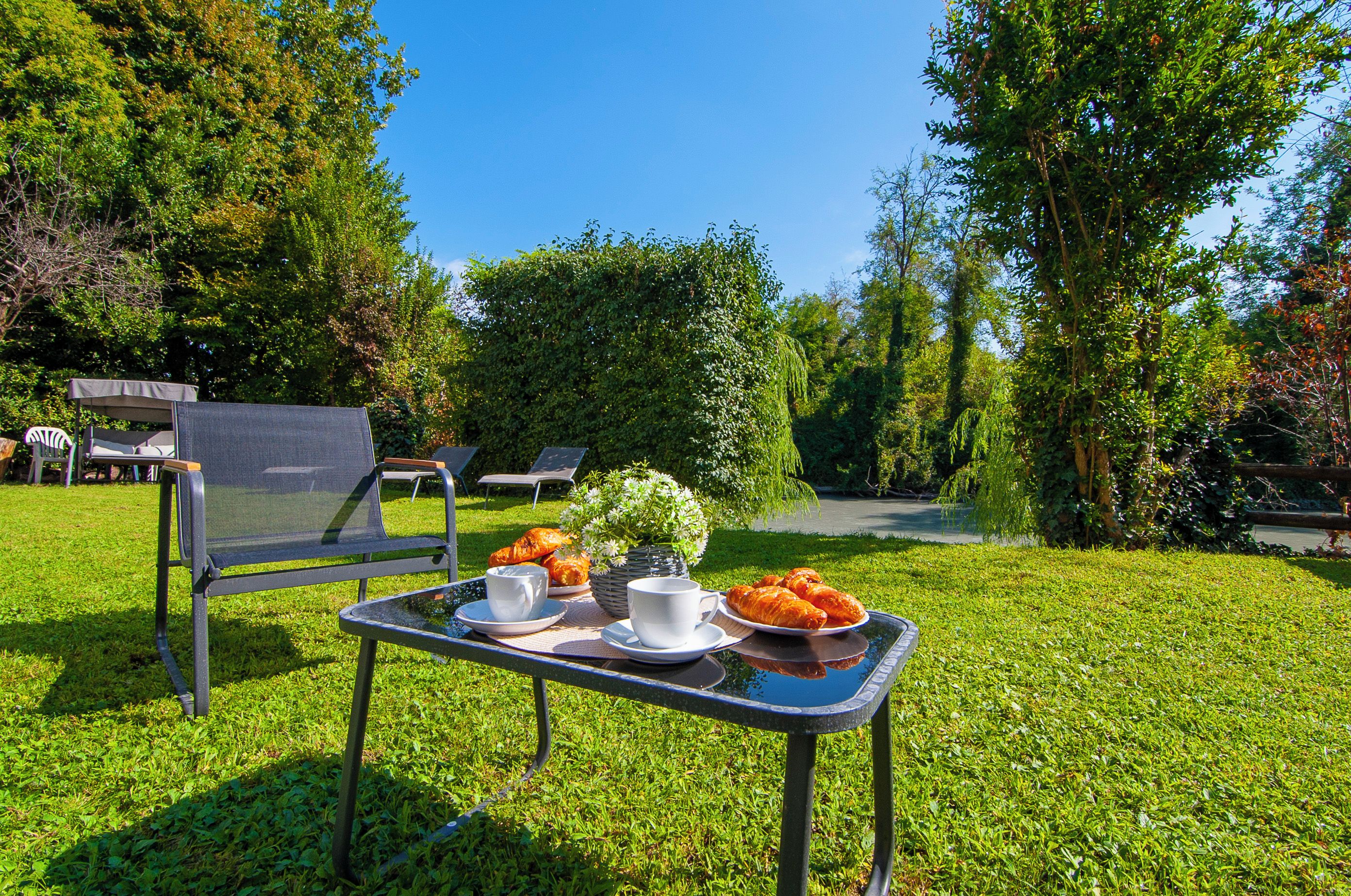 Zona salotto all'aperto con tavolo apparecchiato per un pasto, sedie e vista su un giardino.
