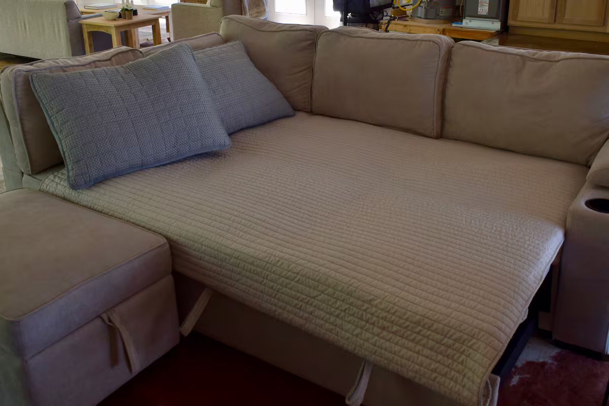 A beige sofa with a quilted cover and two pillows, a matching ottoman, and a small table with a lamp.