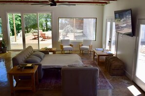 A spacious room with a large bed, a seating area with chairs, a television mounted on the wall, and a view of the outdoors through large windows. - The Studio (Denison)