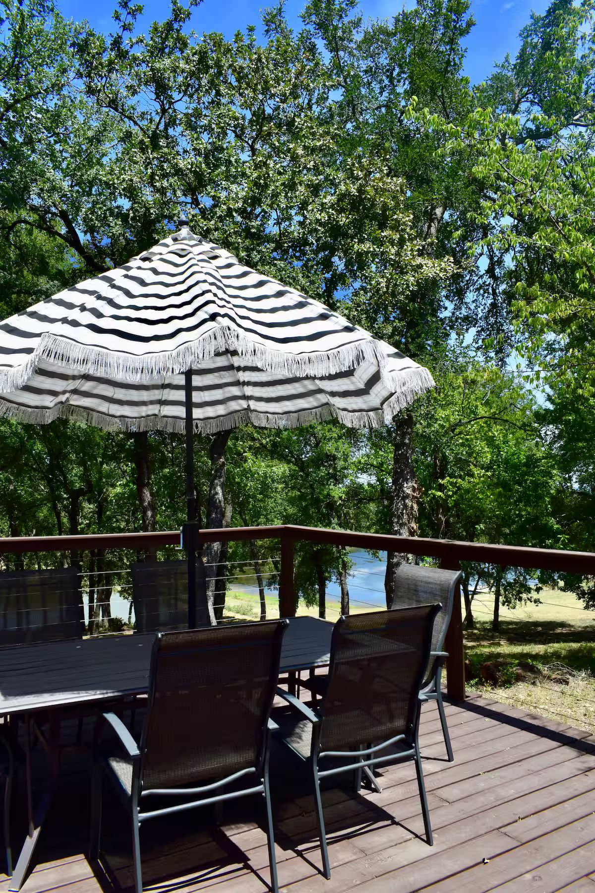 Ein Essbereich im Freien mit Tisch und Stühlen, einem gestreiften Sonnenschirm und Blick auf Bäume und ein Gewässer.