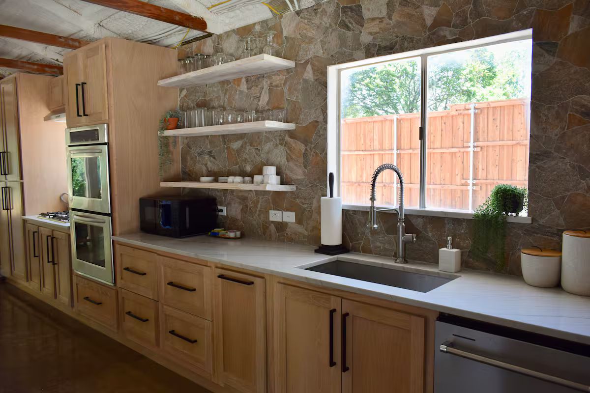 A kitchen with stone walls, wooden cabinets, a stainless steel refrigerator, a microwave, and a window with a view of a wooden fence.