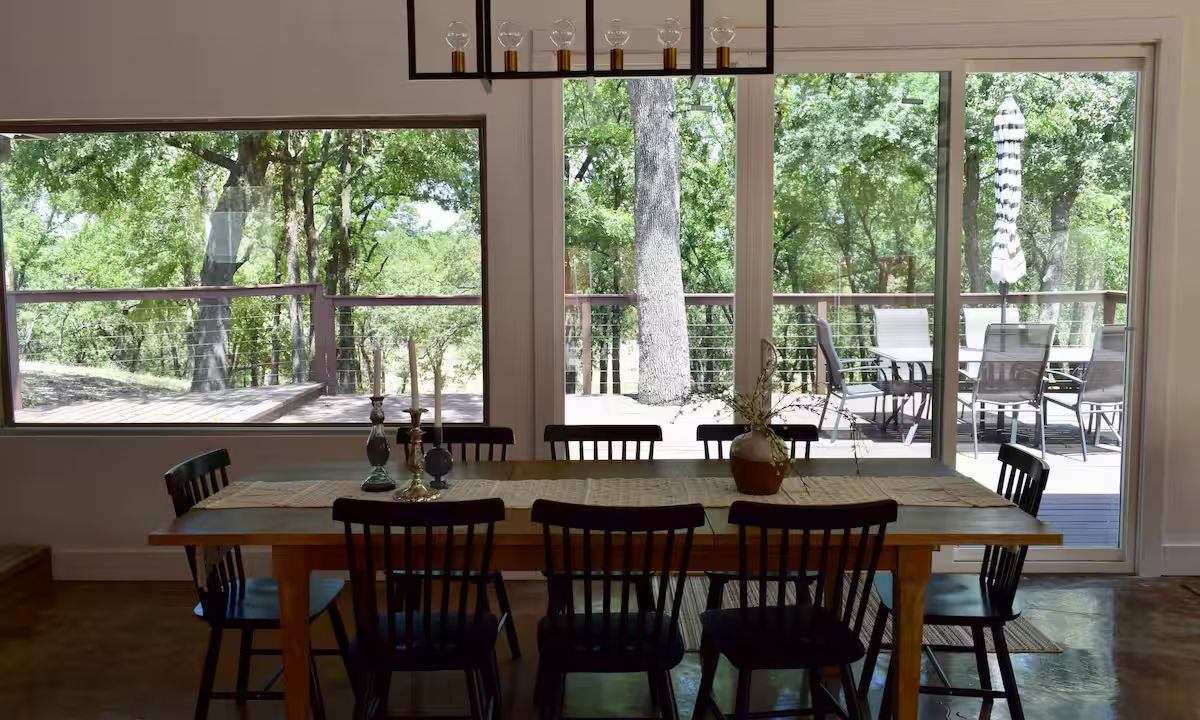 A dining area with a large table and chairs, large windows with a view of trees, and a ceiling light fixture.