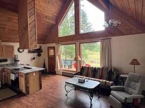 A spacious living area with a wooden ceiling, a kitchen with a central island, and a large window offering a view of the outdoors. - Cabin on 23 acres borders Nat’l forest; close to Blackfoot River, CDT, Lincoln. (Lincoln)