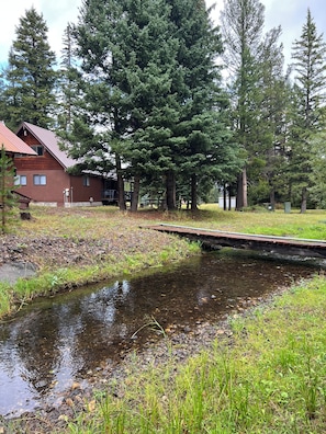Un puente de madera cruzando un arroyo, rodeado de hierba y árboles, con un edificio al fondo.