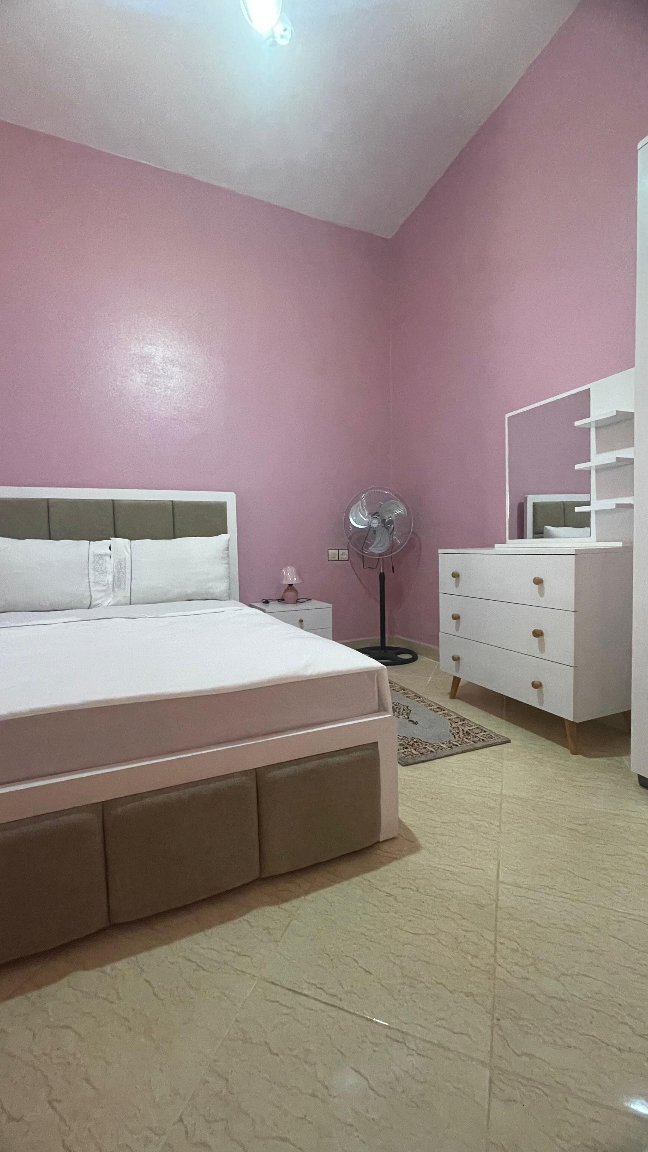 A bedroom with a bed, a bedside table, a lamp, a fan, a dresser, and a mirror.