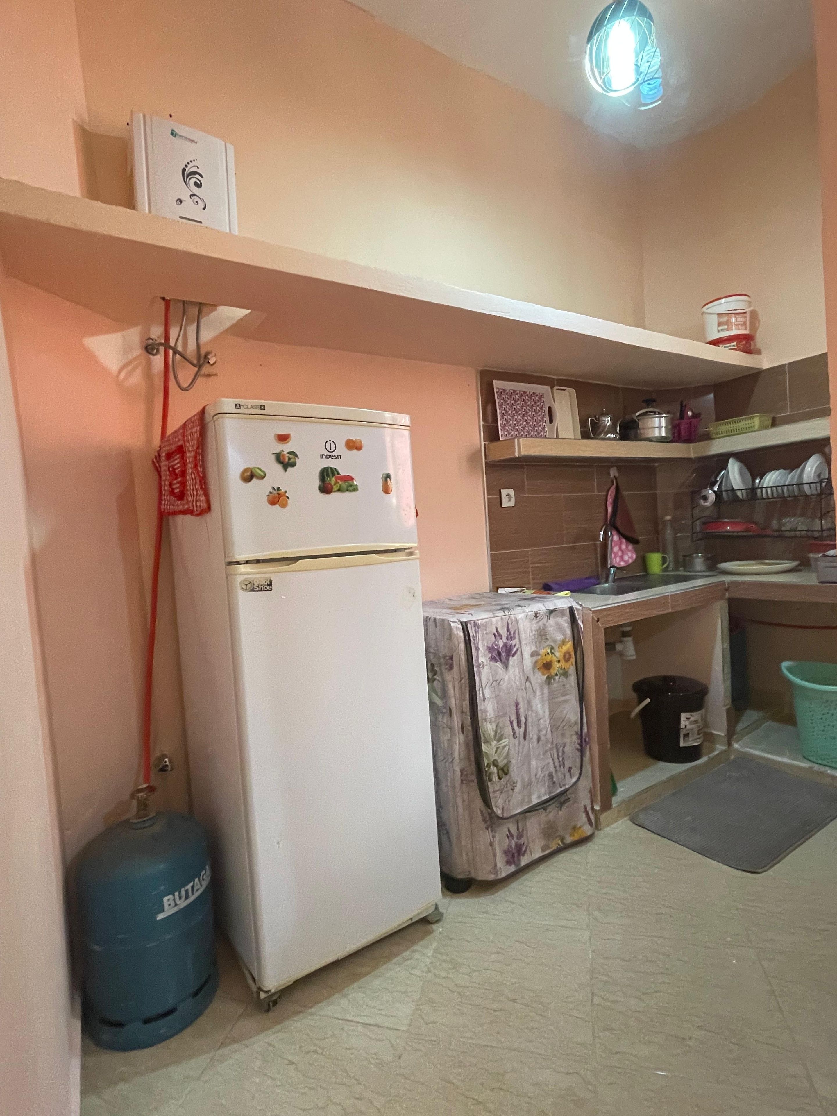 Cocina compacta con refrigerador, cilindro de gas, pileta y un estante con utensilios de cocina.