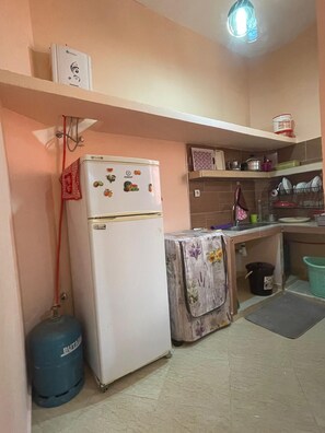 Eine kleine Küche mit Kühlschrank, Gasflasche, Spüle und einem Regal mit Geschirr.