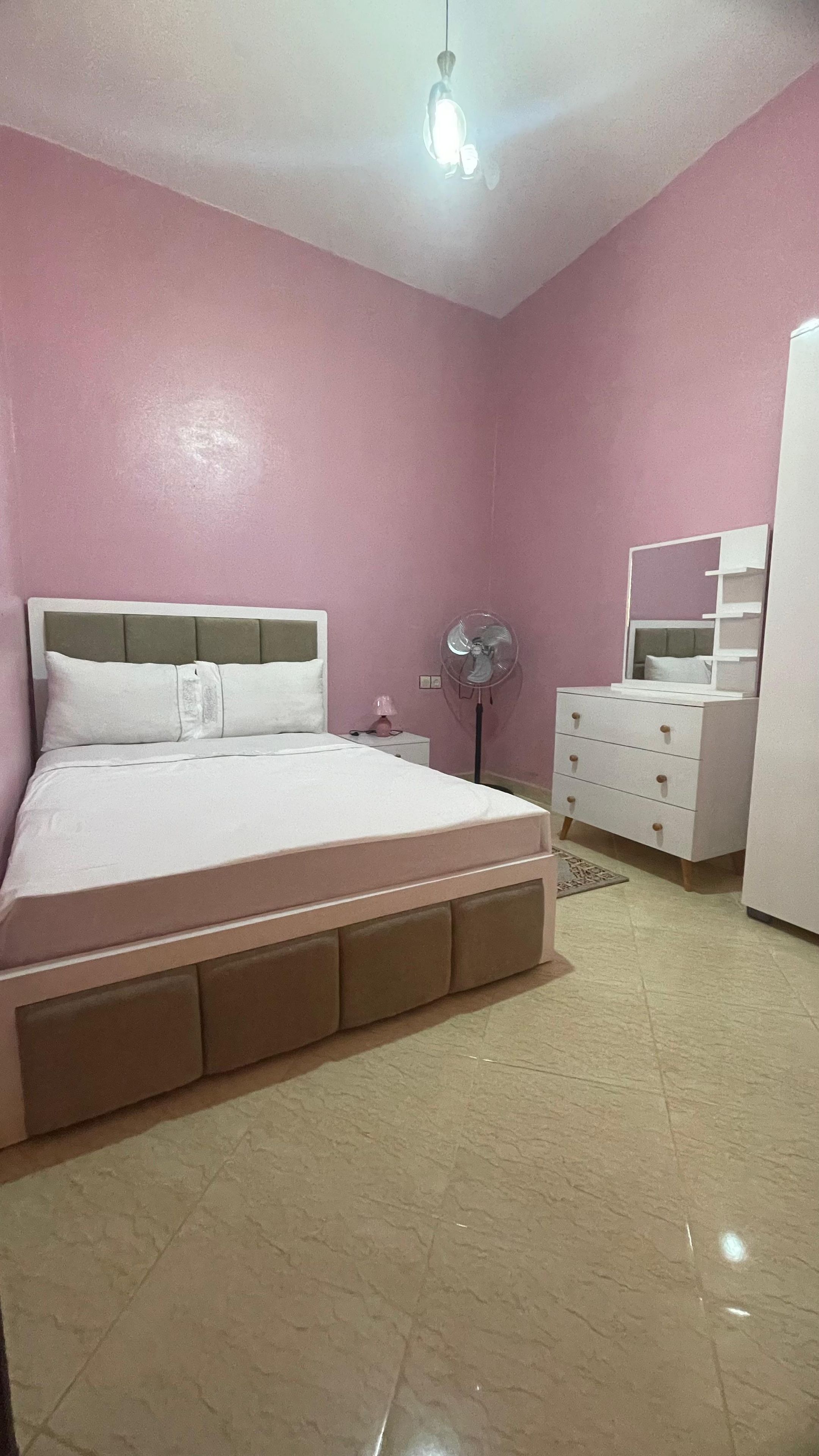Een slaapkamer met een bed, nachtkastje, lamp, ventilator, kapstok en een spiegel.