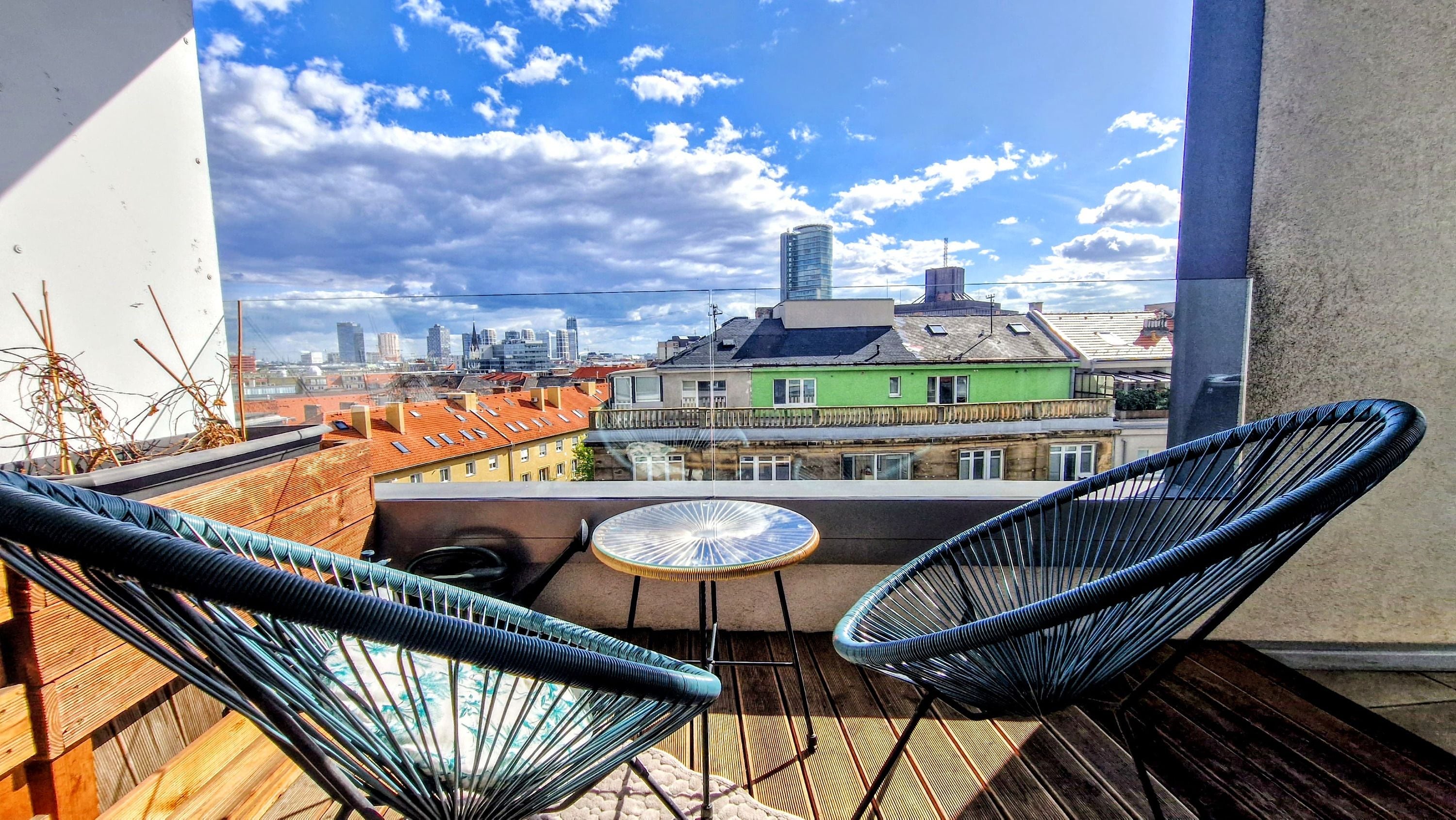 A balcony with two wicker chairs, a small table, and a view of a cityscape with buildings and a clear sky.