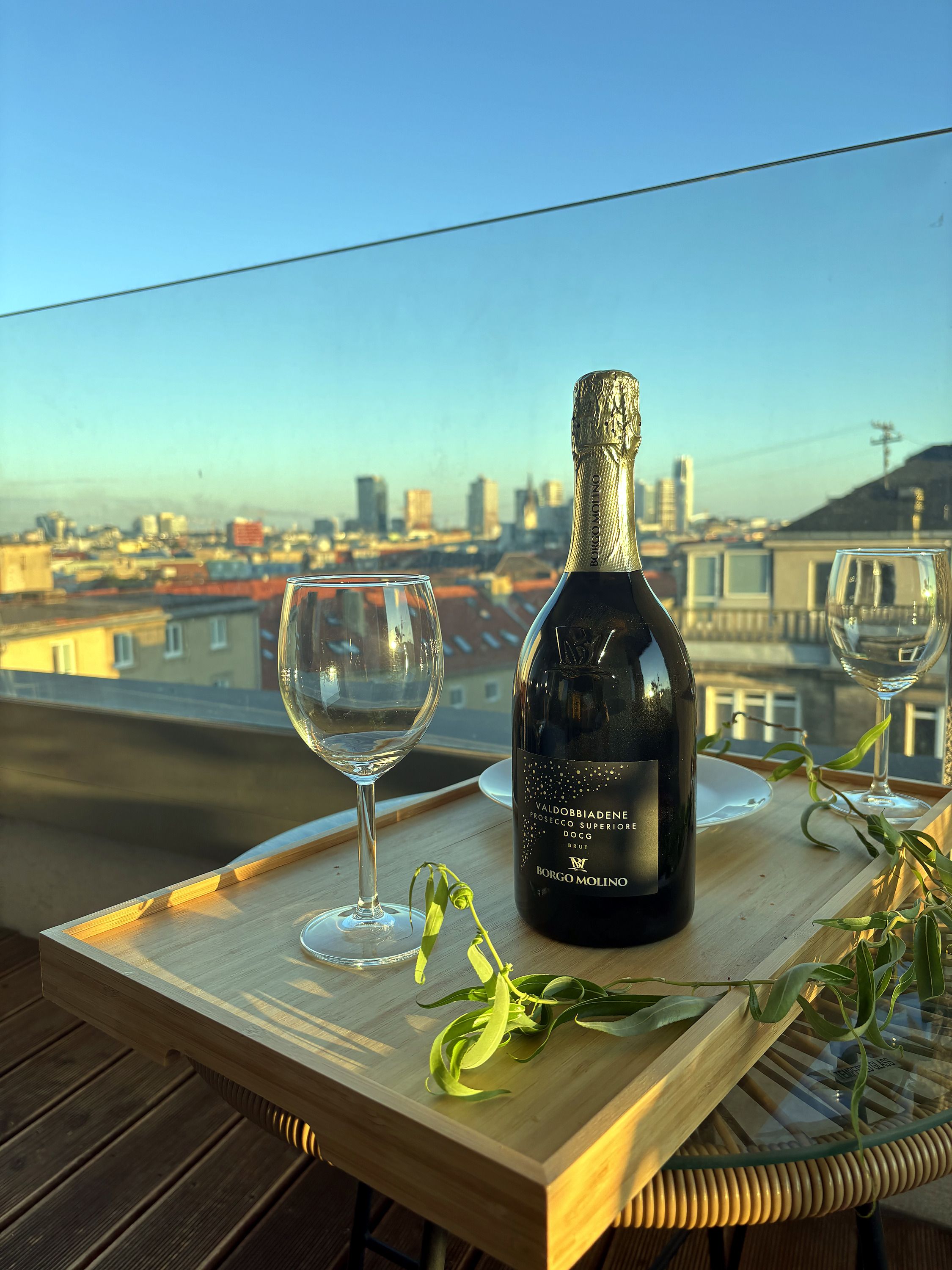 A bottle of wine and two glasses on a wooden tray with a city view in the background.