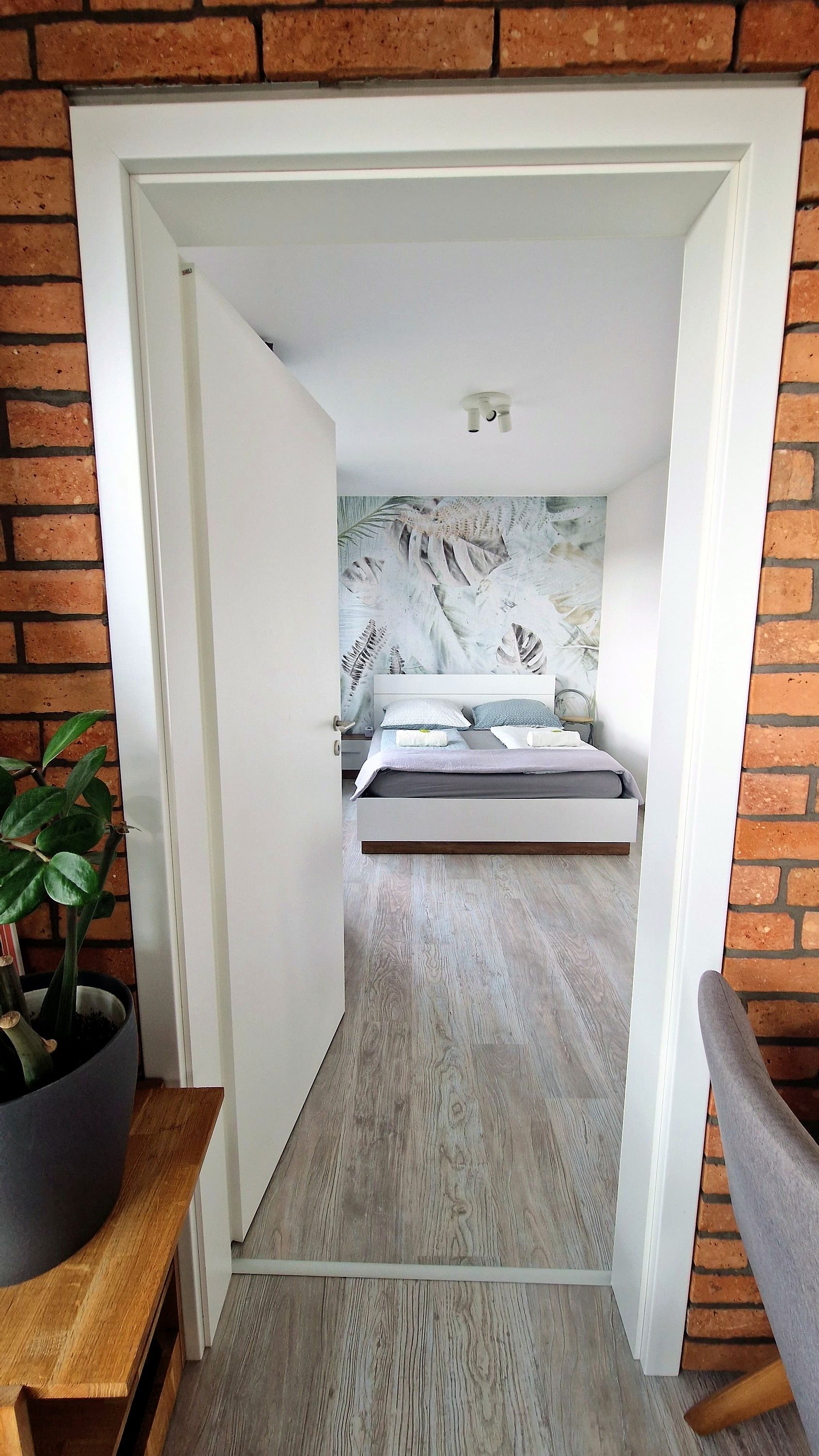 A bedroom with a bed, a nightstand, a potted plant, and a brick wall.