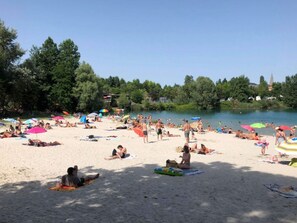 Ein überfüllter Strand mit badenden und sonnenden Personen, umgeben von Bäumen und einem Gewässer.