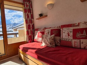 Ein Zimmer mit einem Bett, einem Fenster mit Bergblick, einer an der Wand befestigten Ablage mit einer Pflanze und einem roten und weißen Kissen mit Muster.