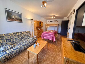 Ein gemütliches Wohnzimmer mit einer gemusterten Couch, einem Holztisch, einem Etagenbett und einem Esstisch für zwei Personen.