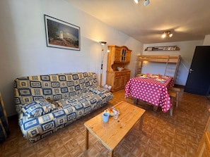 Ein möbliertes Zimmer mit einer Couch, einem Tisch, einem Etagenbett, einem Schrank und einem an der Wand hängenden Bild.