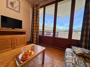 Ein Zimmer mit Fernseher auf einer Holzkabine, einem Esstisch mit einer Schüssel Essen und Besteck sowie einer Couch mit gemusterten Kissen.